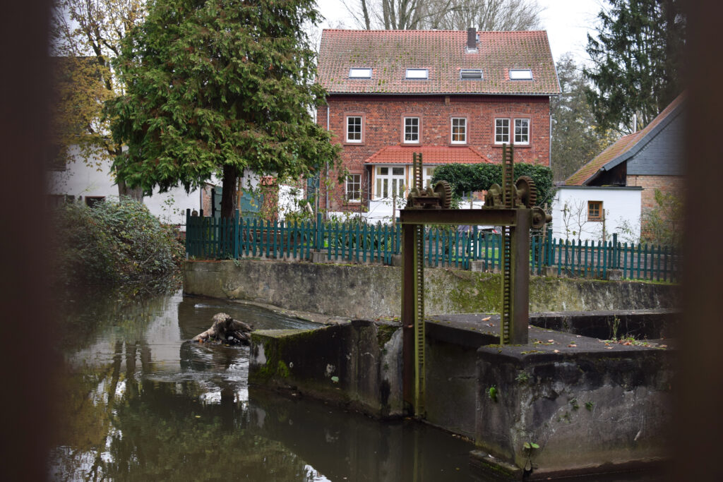 Blick auf ein Wehr in Dieburg, neben dem die leerstehende Mörsmühle steht. Im Hintergrund sind Nadelbäume sowie ein Haus mit roten Ziegelsteinen und weißen Fensterfassaden zu sehen.